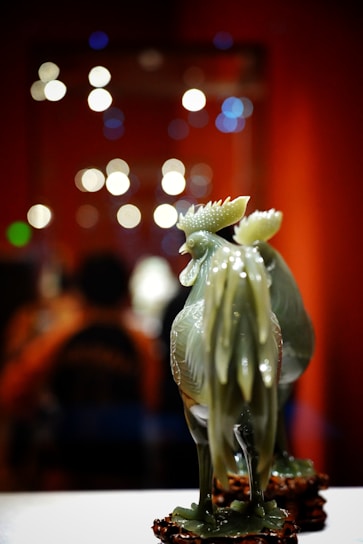 A decorative jade rooster figurine with detailed sculpting stands prominently in the foreground. Its intricate layers reflect light, casting a glossy finish. In the background, a warm bokeh effect with circular light orbs creates a soft, ambient atmosphere, highlighting deep red and orange hues.