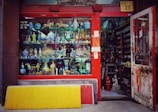 A vibrant storefront showcasing an array of decorative items and antiques, including colorful ceramic vases, figurines, and sculptures. The shop has a red frame around the window and door, with various traditional ornaments hanging from the door. The interior appears densely packed with more collectibles and curiosities.