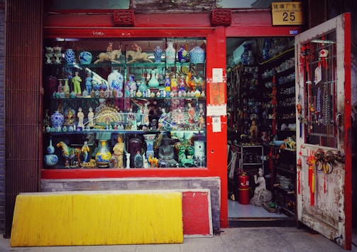 A vibrant storefront showcasing an array of decorative items and antiques, including colorful ceramic vases, figurines, and sculptures. The shop has a red frame around the window and door, with various traditional ornaments hanging from the door. The interior appears densely packed with more collectibles and curiosities.