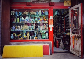 A vibrant storefront showcasing an array of decorative items and antiques, including colorful ceramic vases, figurines, and sculptures. The shop has a red frame around the window and door, with various traditional ornaments hanging from the door. The interior appears densely packed with more collectibles and curiosities.