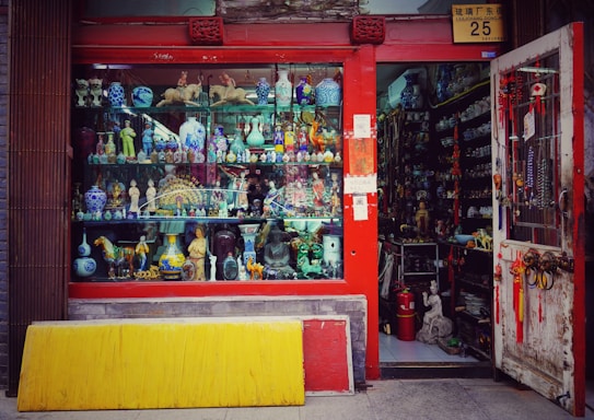 A vibrant storefront showcasing an array of decorative items and antiques, including colorful ceramic vases, figurines, and sculptures. The shop has a red frame around the window and door, with various traditional ornaments hanging from the door. The interior appears densely packed with more collectibles and curiosities.