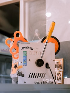 A machine with the brand name 'Jackarmy' is visible. It has a white and silver body with a lever that has a yellow and black handle. There is an orange component attached to the machine, possibly a spool or wheel. Nearby, there are some photo stickers attached to the machine.