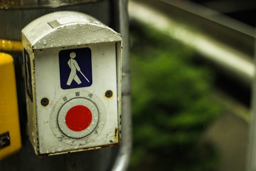 A close-up view of a pedestrian crosswalk button designed for accessibility, featuring a symbol of a person with a cane. The button is metallic with a red circular press area, and is covered in water droplets, indicating recent rain. The background is slightly blurred with greenery visible.