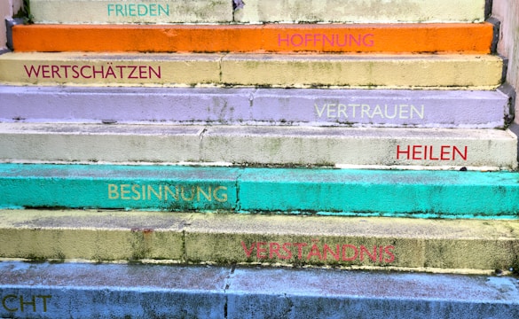 A series of colorful steps with motivational words in German. The steps are painted in various pastel shades including orange, yellow, lavender, turquoise, and green. Words like 'FRIEDEN', 'HOFFNUNG', 'VERTRAUEN', and 'VERSTÄNDNIS' are written on each step, adding an uplifting and positive theme.