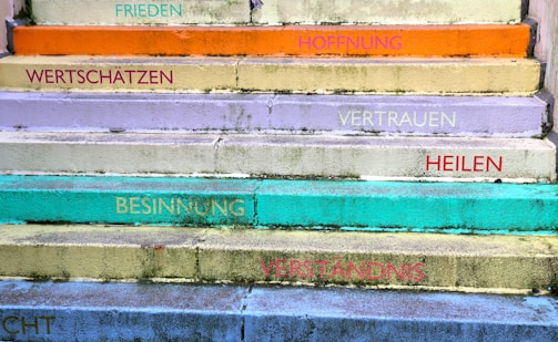 A series of colorful steps with motivational words in German. The steps are painted in various pastel shades including orange, yellow, lavender, turquoise, and green. Words like 'FRIEDEN', 'HOFFNUNG', 'VERTRAUEN', and 'VERSTÄNDNIS' are written on each step, adding an uplifting and positive theme.