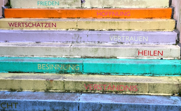 A series of colorful steps with motivational words in German. The steps are painted in various pastel shades including orange, yellow, lavender, turquoise, and green. Words like 'FRIEDEN', 'HOFFNUNG', 'VERTRAUEN', and 'VERSTÄNDNIS' are written on each step, adding an uplifting and positive theme.