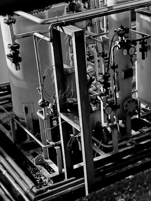 A complex arrangement of industrial pipes, valves, and gauges set against large cylindrical containers. The metallic structure is surrounded by intertwined wires and supported by a framework of beams and rails. The image is monochromatic, highlighting textures and contrasts within the equipment.