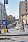 A lively urban scene features a canal flanked by residential buildings and a bustling walkway. People are strolling along the path, some carrying bags or pushing carts. Overhead, street signs and utility wires intersect while the sky is clear with a few clouds.