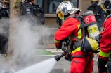 Firefighters wearing red protective suits and metallic helmets are using a hose to spray water towards thick white smoke. They are equipped with breathing apparatus and additional gear. In the background, police officers in dark uniforms and helmets stand by a building.