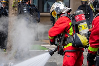 Firefighters wearing red protective suits and metallic helmets are using a hose to spray water towards thick white smoke. They are equipped with breathing apparatus and additional gear. In the background, police officers in dark uniforms and helmets stand by a building.