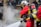 Firefighters wearing red protective suits and metallic helmets are using a hose to spray water towards thick white smoke. They are equipped with breathing apparatus and additional gear. In the background, police officers in dark uniforms and helmets stand by a building.