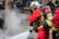Firefighters wearing red protective suits and metallic helmets are using a hose to spray water towards thick white smoke. They are equipped with breathing apparatus and additional gear. In the background, police officers in dark uniforms and helmets stand by a building.