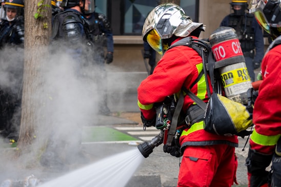 Firefighters wearing red protective suits and metallic helmets are using a hose to spray water towards thick white smoke. They are equipped with breathing apparatus and additional gear. In the background, police officers in dark uniforms and helmets stand by a building.