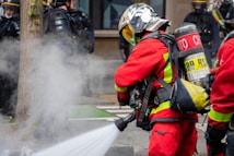 Firefighters wearing red protective suits and metallic helmets are using a hose to spray water towards thick white smoke. They are equipped with breathing apparatus and additional gear. In the background, police officers in dark uniforms and helmets stand by a building.