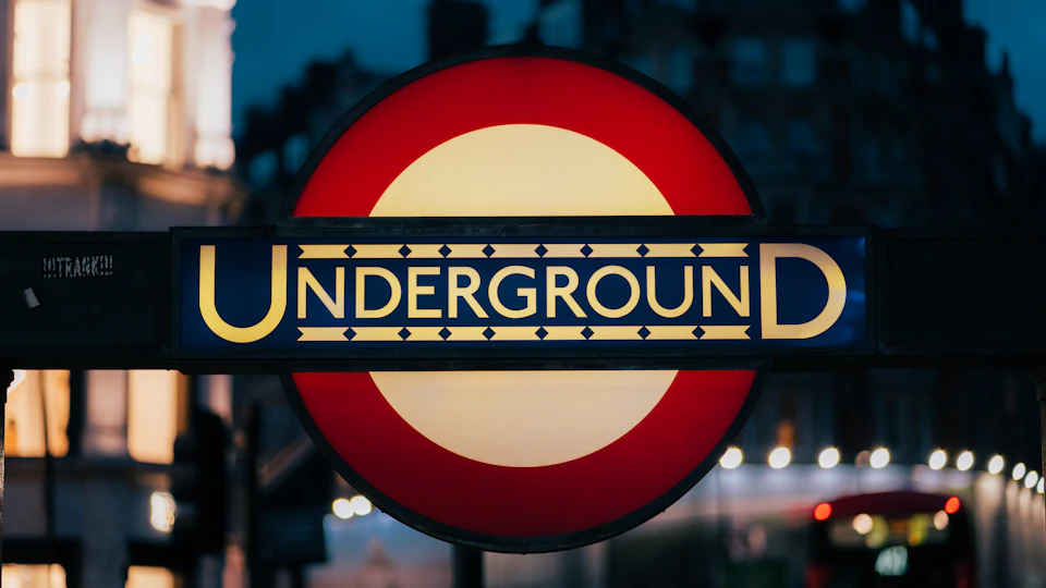 The image shows a round sign for the Underground, illuminated with a warm yellow glow against a dark background. The sign is prominently displayed on a pole and features the words "Underground" written in bold black letters at the top, followed by a smaller white circle containing the word "D" below it. Below this, there's a larger red circle enclosing the word "U". In the blurred background, you can see buildings and streetlights, suggesting that the sign is located in an urban setting with city lights illuminating the area around it.