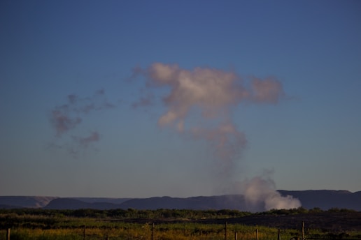 A scenic landscape with a clear blue sky and a few wispy clouds. In the foreground, lush greenery is visible, while hills or mountains can be seen in the distance. A plume of smoke or mist rises from behind the hills.
