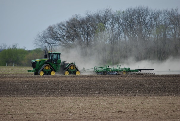A green tractor with caterpillar tracks is plowing a large field, creating clouds of dust. The field appears expansive and freshly tilled, ready for planting. In the background, there are leafless trees and some greenery, indicating early spring.