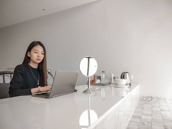 A woman is seated at a sleek, white, marble-top table, focusing intently on a laptop. The setting appears to be minimalist and modern, with a small round mirror, a teapot, and a few other items like bottles and cups neatly arranged on the table.