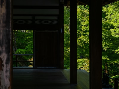 A tranquil wooden terrace with dark wooden pillars overlooks a lush green forest. The light filtering through the trees casts gentle shadows on the wooden floor, creating a serene and peaceful atmosphere.