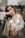 A woman with long dark hair is wearing a white bra and an unbuttoned beige shirt. She is gently resting one hand on her pregnant belly, while her other arm is raised, partially covering her face. The background is blurred, suggesting an indoor setting with some outdoor view.