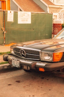 A classic Mercedes-Benz car is parked on the side of the street. The car's front grille and emblem are prominently visible, and it has an old-fashioned license plate. The background includes part of a house and a green construction fence with posted notices.