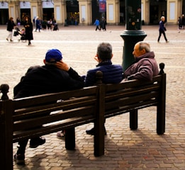 Three elderly people sit on a bench in a spacious cobblestone square. They appear to be casually chatting, with one wearing a blue cap and another with a visible gray head. Several passersby walk in the background near historic-looking buildings with arched doorways. The lighting creates a warm, relaxed atmosphere.