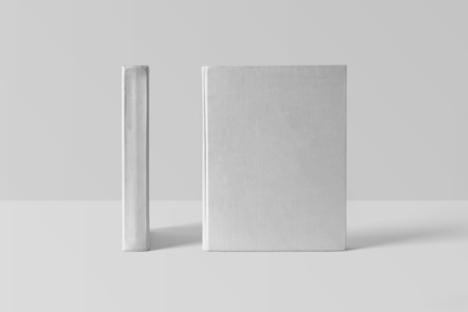 Two plain, hardcover books stand upright against a minimalist background. One book is positioned with its spine facing the front, while the other is showing the front cover.