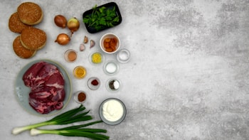 A variety of cooking ingredients are neatly arranged on a gray surface. Elements include raw meat on a plate, sesame seed burger buns, whole onions, cloves of garlic, green onions, spices in small dishes, and fresh parsley in a black container. There are also small bowls containing liquids and powders.