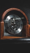 An antique wooden clock with visible inner mechanisms and Roman numeral markings on its face. The clock is set against a dark background, highlighting its intricate details.