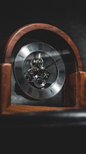 An antique wooden clock with visible inner mechanisms and Roman numeral markings on its face. The clock is set against a dark background, highlighting its intricate details.