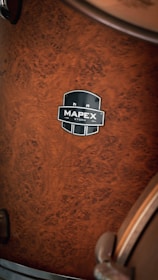 A close-up view of a wooden drum with a rich, brown burl pattern. The drum features a metallic badge with the brand name 'Mapex' and model 'Storm'. The texture of the wood is prominent and the lighting highlights the intricate details of the grain.
