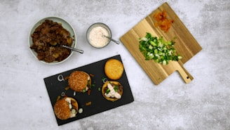 A flatlay featuring a variety of ingredients and prepared food. A black slate plate holds three mini burgers topped with greens and sauces. Adjacent is a wooden cutting board with chopped spring onions and sliced carrots. A bowl of shredded meat sits on the left with a spoon, and a small bowl of creamy dressing is placed near the center.