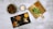 A flatlay featuring a variety of ingredients and prepared food. A black slate plate holds three mini burgers topped with greens and sauces. Adjacent is a wooden cutting board with chopped spring onions and sliced carrots. A bowl of shredded meat sits on the left with a spoon, and a small bowl of creamy dressing is placed near the center.