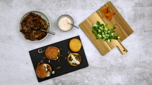 A flatlay featuring a variety of ingredients and prepared food. A black slate plate holds three mini burgers topped with greens and sauces. Adjacent is a wooden cutting board with chopped spring onions and sliced carrots. A bowl of shredded meat sits on the left with a spoon, and a small bowl of creamy dressing is placed near the center.