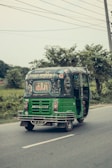A green auto-rickshaw is driving along a road surrounded by greenery. The back of the vehicle is adorned with colorful decorative patterns and text, possibly written in a local language. The sky appears overcast, and there are power lines above.