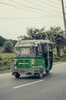 A green auto-rickshaw is driving along a road surrounded by greenery. The back of the vehicle is adorned with colorful decorative patterns and text, possibly written in a local language. The sky appears overcast, and there are power lines above.