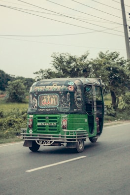 A green auto-rickshaw is driving along a road surrounded by greenery. The back of the vehicle is adorned with colorful decorative patterns and text, possibly written in a local language. The sky appears overcast, and there are power lines above.