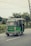 A green auto-rickshaw is driving along a road surrounded by greenery. The back of the vehicle is adorned with colorful decorative patterns and text, possibly written in a local language. The sky appears overcast, and there are power lines above.