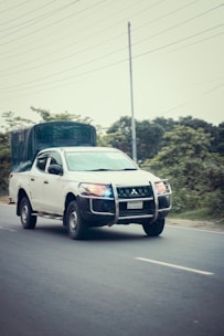A white pickup truck with a protective grille and a covered cargo area is driving on a paved road. The vehicle has blue and red lights signaling its possible use in an official or emergency capacity. The background includes greenery and utility poles.