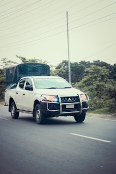A white pickup truck with a protective grille and a covered cargo area is driving on a paved road. The vehicle has blue and red lights signaling its possible use in an official or emergency capacity. The background includes greenery and utility poles.