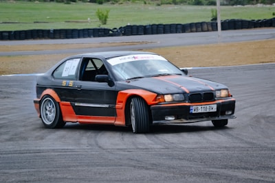 A car is drifting on a racetrack, with smoke visible from its tires. The car is a black and orange sporty coupe with racing numbers displayed on the side windows. The setting is open, with a backdrop of tire barriers and a grassy area beyond the track.