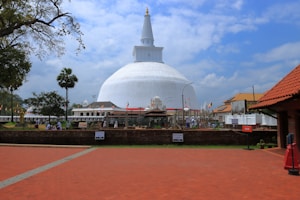 A large white stupa stands majestically under a blue, partly cloudy sky. Surrounding it are people walking and sitting in a courtyard with red brick paths. Greenery and palm trees add a natural element to the scene, while flags and structures indicate a cultural or religious significance.