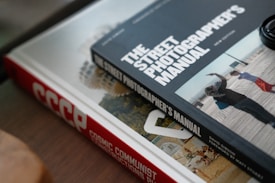 Two books are placed on a surface, one on top of the other. The book on top is titled 'THE STREET PHOTOGRAPHER'S MANUAL' and contains a photo of people walking. The book underneath has a red spine with the title 'COSMIC COMMUNIST CONSTRUCTIONS'. Part of another book is visible under the top book, showing a circular building.