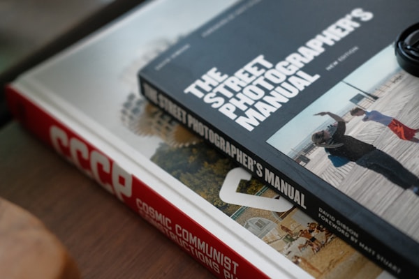 Two books are placed on a surface, one on top of the other. The book on top is titled 'THE STREET PHOTOGRAPHER'S MANUAL' and contains a photo of people walking. The book underneath has a red spine with the title 'COSMIC COMMUNIST CONSTRUCTIONS'. Part of another book is visible under the top book, showing a circular building.