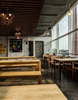The image depicts an empty restaurant with minimalistic wooden furniture and a wall decorated with white and black doodles. The ceiling has multiple lantern-style light fixtures, and large windows on one side allow ample daylight to stream in. A blackboard on one of the walls reads 'Asian Street Food' and displays several menu items. The overall ambiance is modern and relaxed.
