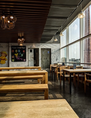 The image depicts an empty restaurant with minimalistic wooden furniture and a wall decorated with white and black doodles. The ceiling has multiple lantern-style light fixtures, and large windows on one side allow ample daylight to stream in. A blackboard on one of the walls reads 'Asian Street Food' and displays several menu items. The overall ambiance is modern and relaxed.