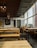 The image depicts an empty restaurant with minimalistic wooden furniture and a wall decorated with white and black doodles. The ceiling has multiple lantern-style light fixtures, and large windows on one side allow ample daylight to stream in. A blackboard on one of the walls reads 'Asian Street Food' and displays several menu items. The overall ambiance is modern and relaxed.