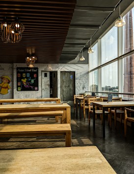 The image depicts an empty restaurant with minimalistic wooden furniture and a wall decorated with white and black doodles. The ceiling has multiple lantern-style light fixtures, and large windows on one side allow ample daylight to stream in. A blackboard on one of the walls reads 'Asian Street Food' and displays several menu items. The overall ambiance is modern and relaxed.