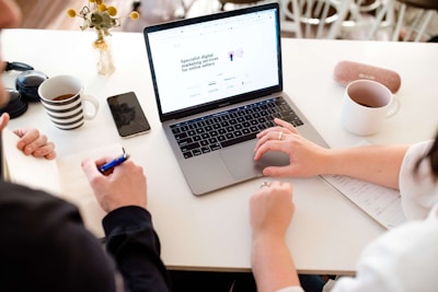 Two people are seated at a white table engaged in a collaborative work session. One person's hands hover over a laptop with a webpage open on its screen, while the other person writes in a notebook. A striped mug, a smartphone, a pair of headphones, another mug, and a vase with flowers are also present on the table.