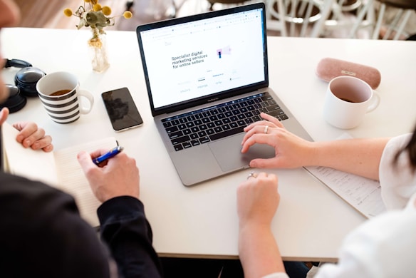 Two people are seated at a white table engaged in a collaborative work session. One person's hands hover over a laptop with a webpage open on its screen, while the other person writes in a notebook. A striped mug, a smartphone, a pair of headphones, another mug, and a vase with flowers are also present on the table.
